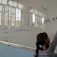 Ein Mädchen schreibt "DANKE HHC" an eine weiße Tafel. Die hellen Fenster des Klassenraums spiegeln sich in der Tafel.